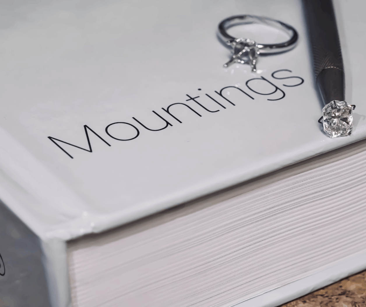 A silver ring mounting with empty prongs and a loose round diamond resting on a white book titled "Mountings" next to a professional jeweler’s tool