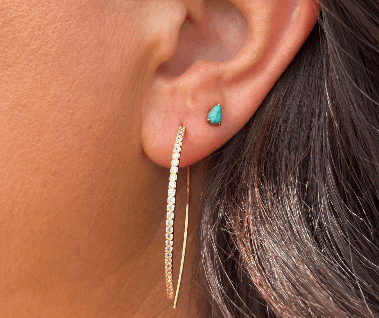 A close-up side view of a person’s ear wearing a small teardrop-shaped turquoise stud earring and a large, thin gold hoop lined with shimmering pavé diamonds