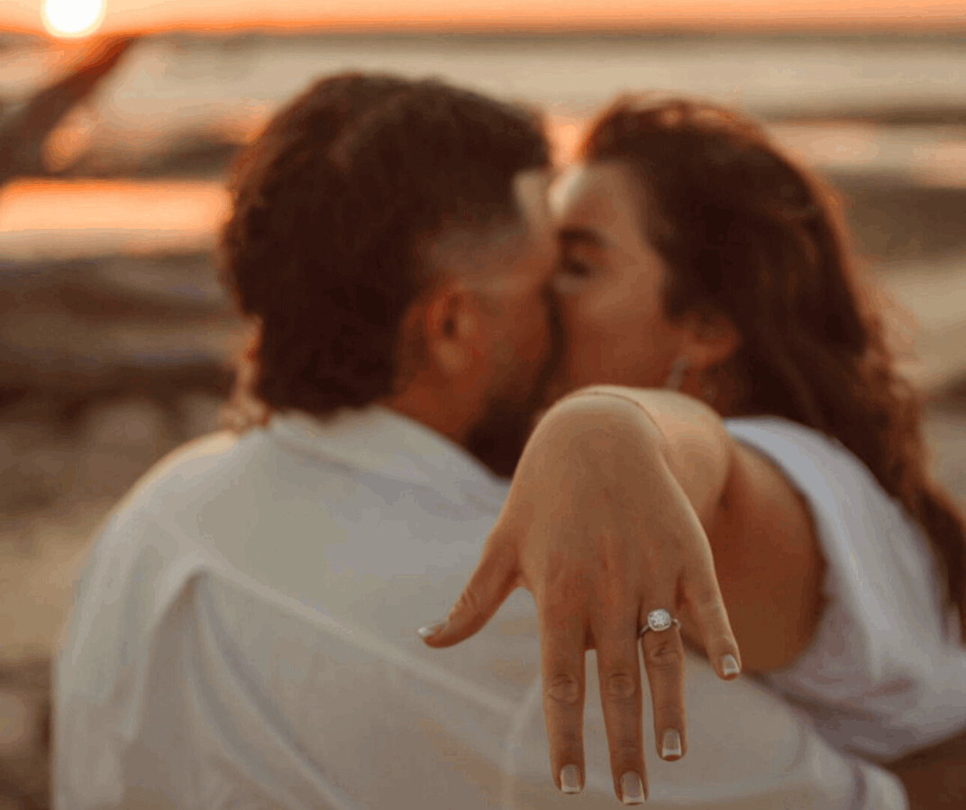 Close-up of a diamond engagement ring on a woman's hand with a couple kissing in the background