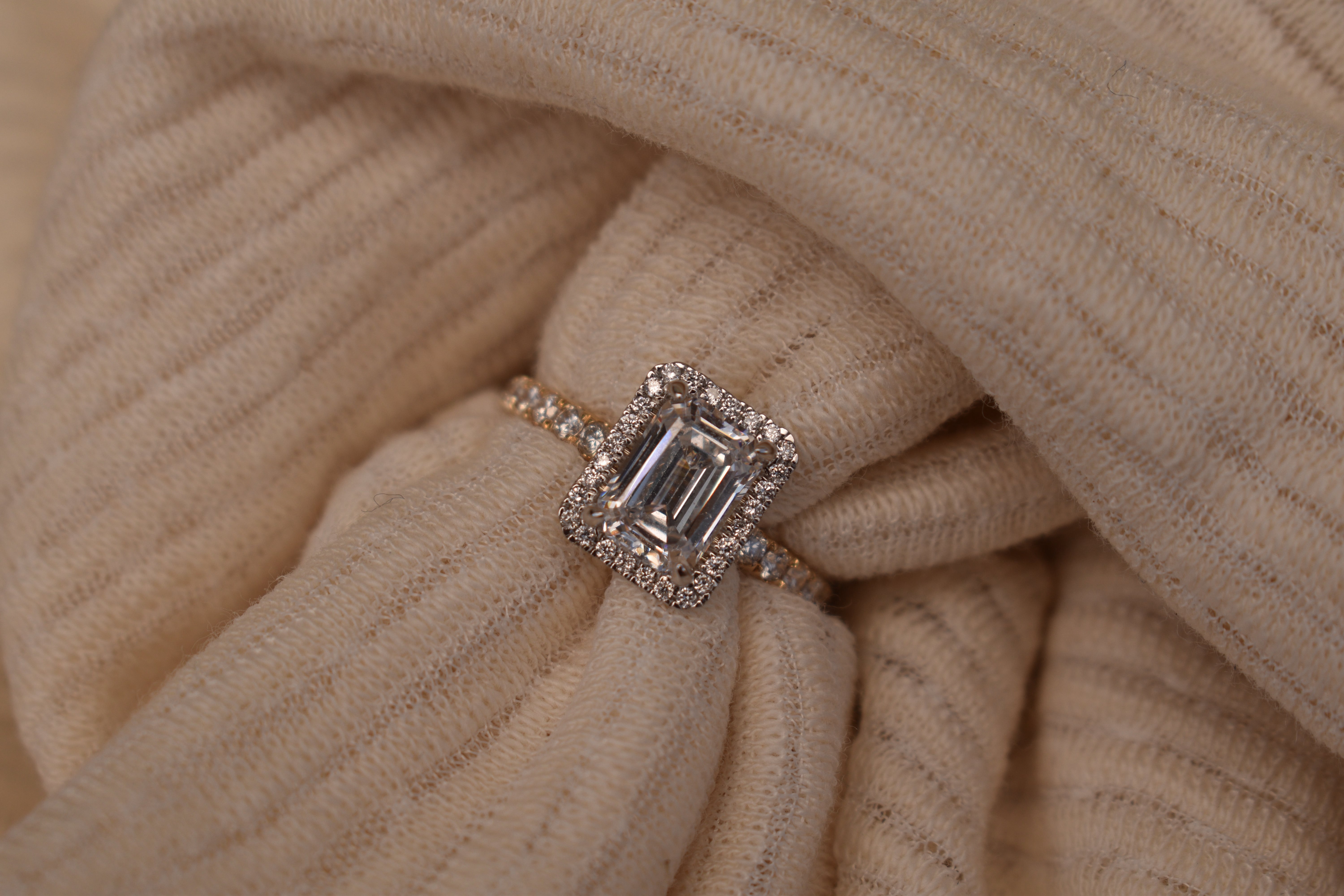 Gold engagement ring featuring a large emerald-cut center diamond with a pavé halo and diamond band, nestled within the folds of a cream ribbed fabric