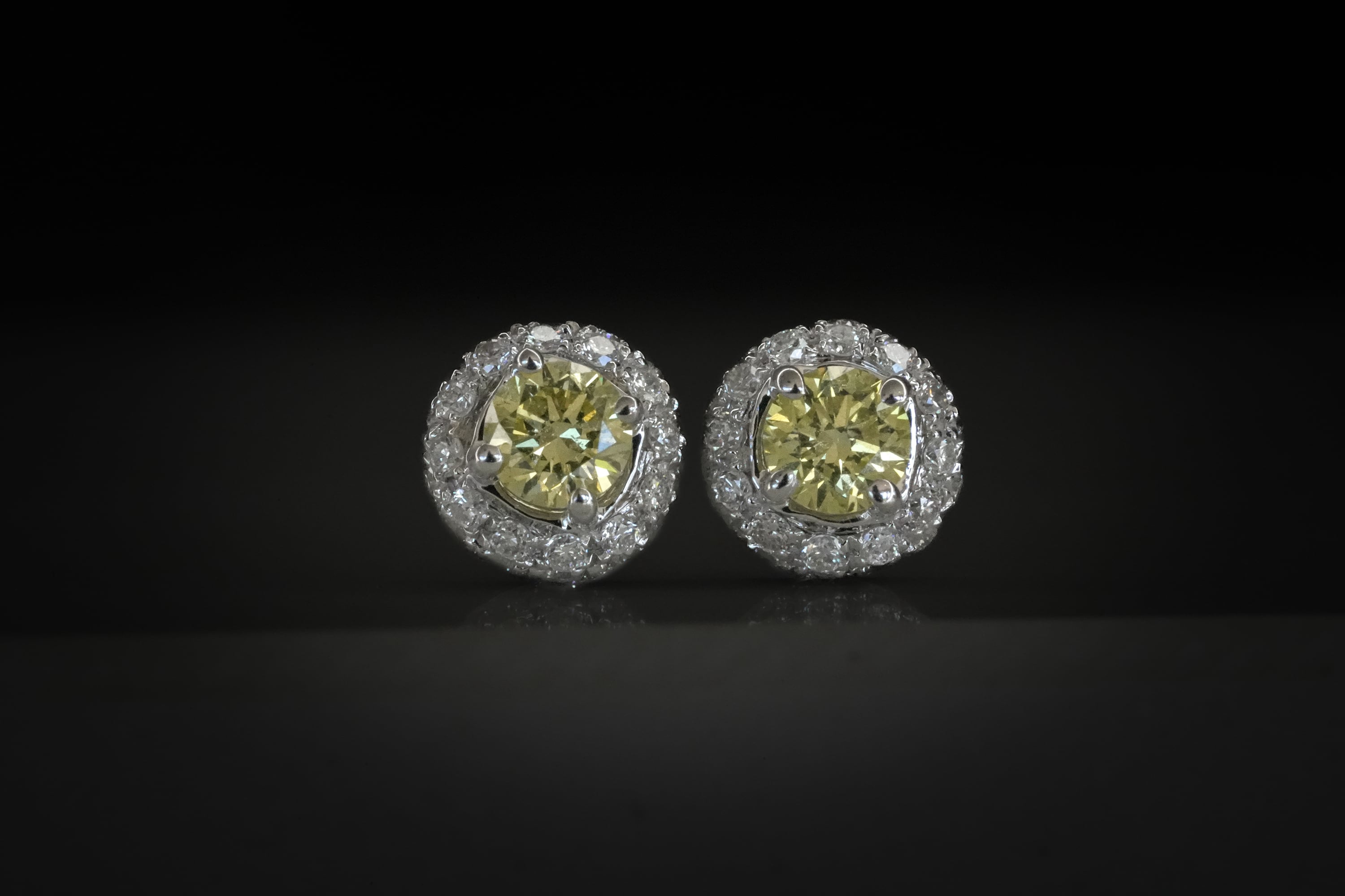 Silver halo stud earrings featuring round yellow diamond center stones surrounded by a border of small white diamonds on a dark reflective surface