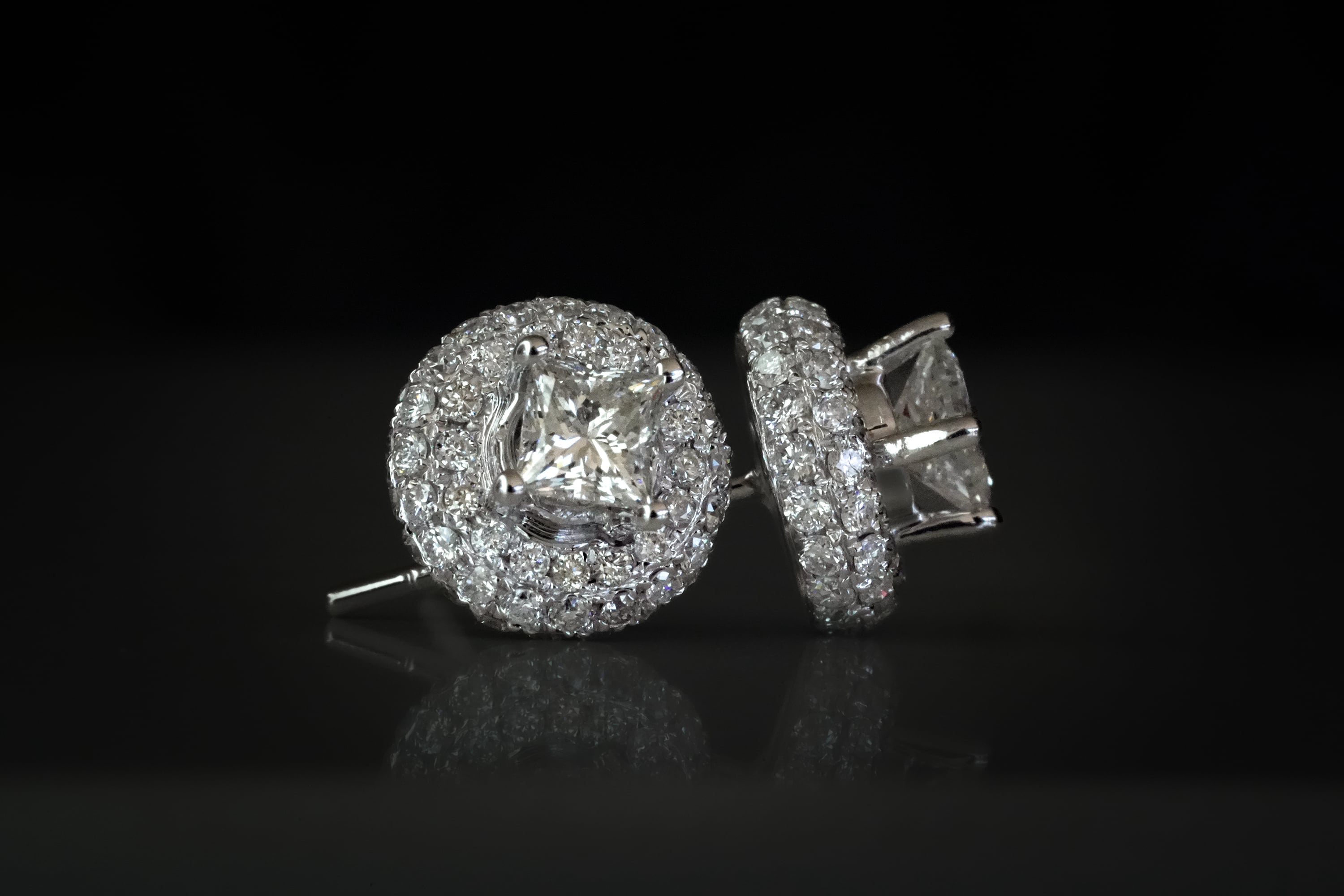 Silver halo stud earrings featuring a princess-cut center diamond surrounded by two rows of pave-set round diamonds on a dark reflective surface