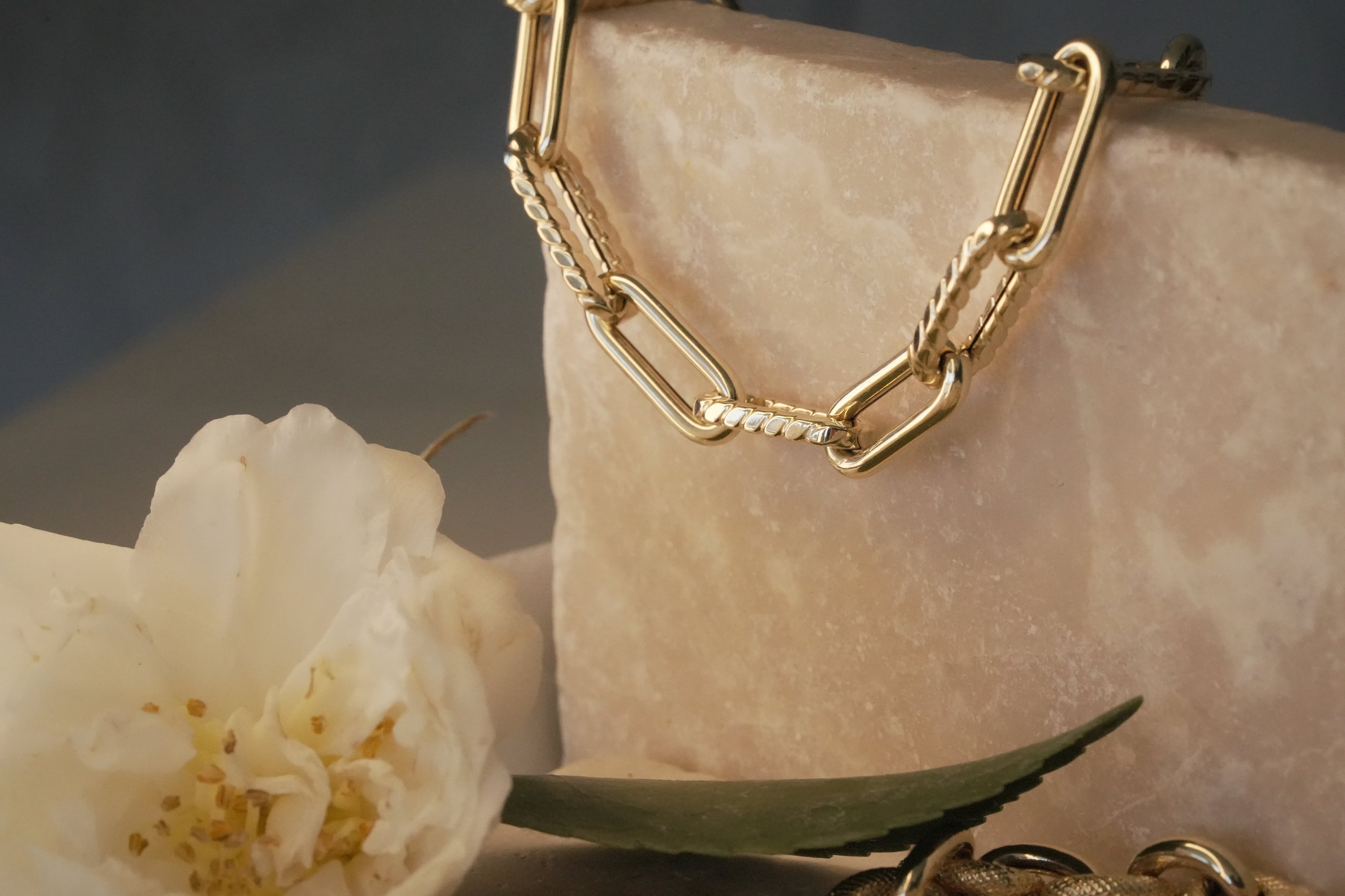 A close-up of a 14k yellow gold paperclip link necklace, featuring alternating smooth and textured rope-style links, draped over a beige marble block next to a white flower