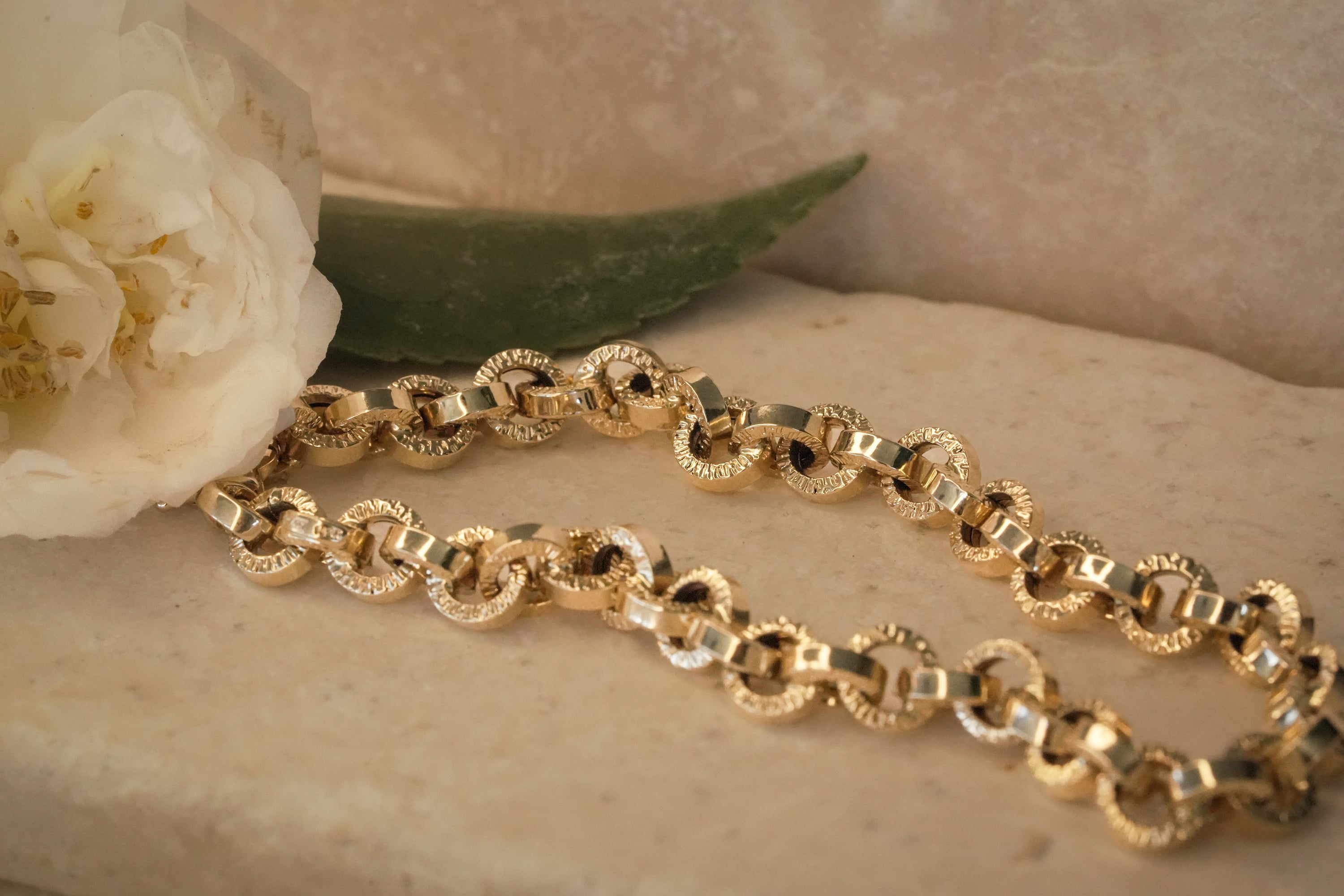 Gold chain bracelet with alternating polished and textured round links, displayed on a stone surface next to a white flower and green leaf