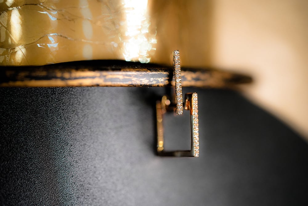 Gold geometric hoop earrings featuring a modern square design accented with a row of small pave diamonds, suspended against a dark textured surface